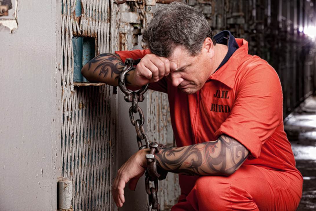 Man in orange jumpsuit with "JAIL BIRD" printed on the back sits in jail, looking distraught. His wrists are shackled with heavy chains.